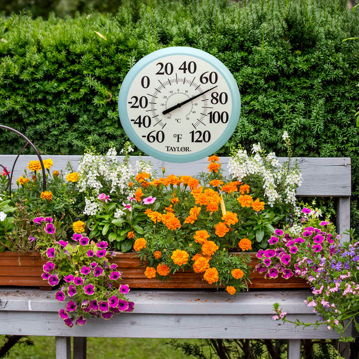 13.25 inch outdoor thermometer with a light blue rim set in a garden among green shrubs and colorful flowers