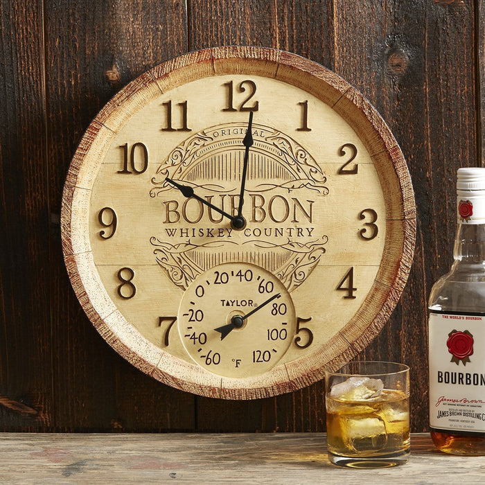 Rustic 14 inch Bourbon Barrel Clock with thermometer mounted on a dark wooden wall beside a whiskey bottle.
