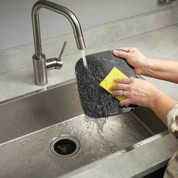Hands washing the black marble digital kitchen scale under a running faucet in a sink
