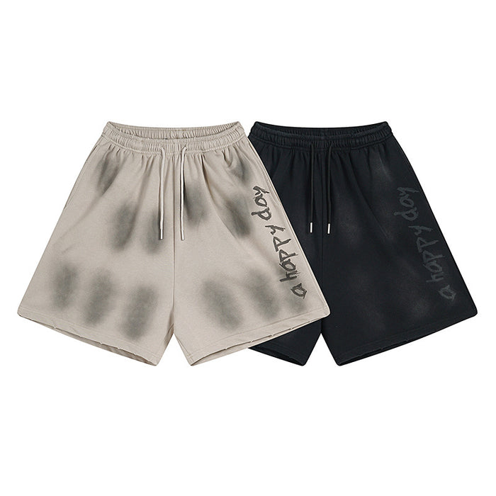 Front view of washed beige and black relaxed-fit shorts with elastic waist, drawstrings, and side graphic