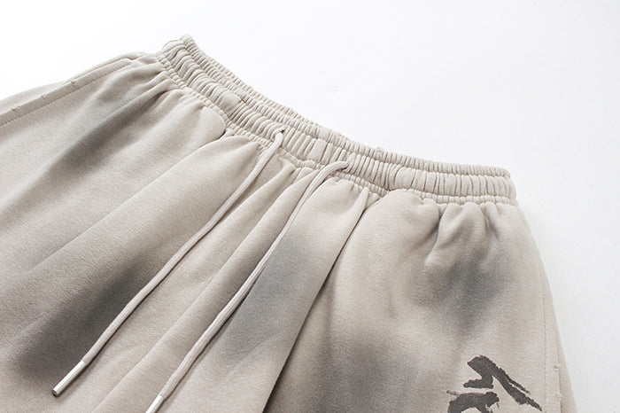 Close-up of beige shorts showing gathered elastic waistband, drawstrings, washed shading, and partial graphic