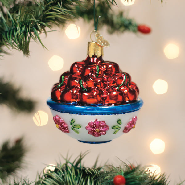 A glass Christmas tree ornament shaped like a bowl of shiny red cherries with green stems, decorated with pink flowers and green leaves, hanging on a tree branch with blurred lights in the background.