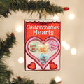 A glass ornament shaped like a candy box labeled Conversation Hearts, featuring pastel heart candies with messages like KISS ME, I LUV YOU, HUG ME, and BE MINE, hanging on a Christmas tree.