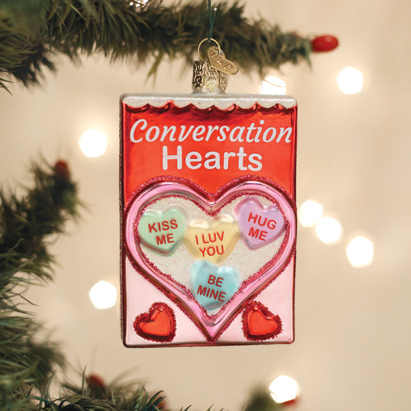 A glass ornament shaped like a candy box labeled Conversation Hearts, featuring pastel heart candies with messages like KISS ME, I LUV YOU, HUG ME, and BE MINE, hanging on a Christmas tree.