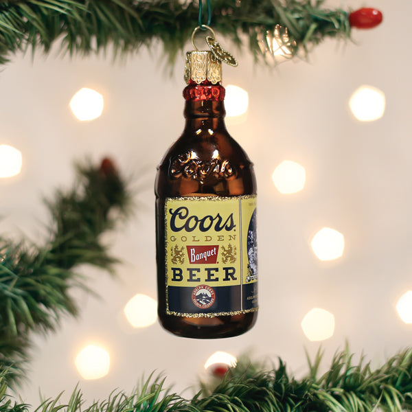 A Christmas tree ornament shaped like a brown Coors Banquet beer bottle hangs from a branch, with blurred holiday lights in the background.