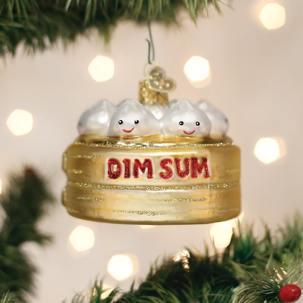A Christmas tree ornament shaped like a bamboo steamer with smiling dumplings inside, labeled DIM SUM in red glitter, hangs among green branches with blurred lights in the background.
