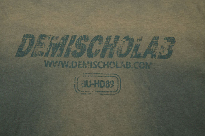 Close-up of the olive shirt chest graphic showing distressed lettering, website text and a rounded outlined emblem