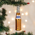 A glass ornament shaped like a classic Fanta soda bottle hangs on a green Christmas tree branch, with soft white lights blurred in the background.