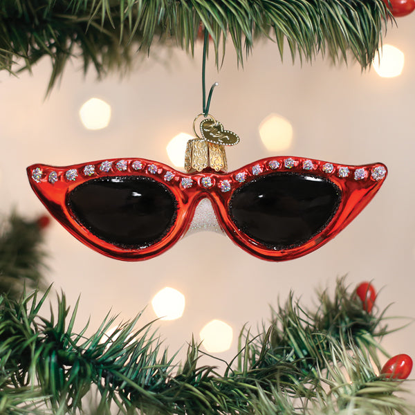 A Christmas tree ornament shaped like red cat-eye sunglasses with rhinestone details, hanging on a green tree branch with blurred holiday lights in the background.