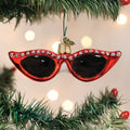A Christmas tree ornament shaped like red cat-eye sunglasses with rhinestone details, hanging on a green tree branch with blurred holiday lights in the background.