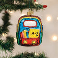A colorful, glass ornament shaped like a school backpack with pencils, books, and decorations hangs on a Christmas tree with blurred white lights in the background.