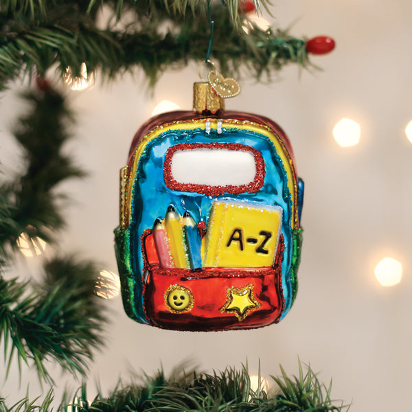 A colorful, glass ornament shaped like a school backpack with pencils, books, and decorations hangs on a Christmas tree with blurred white lights in the background.