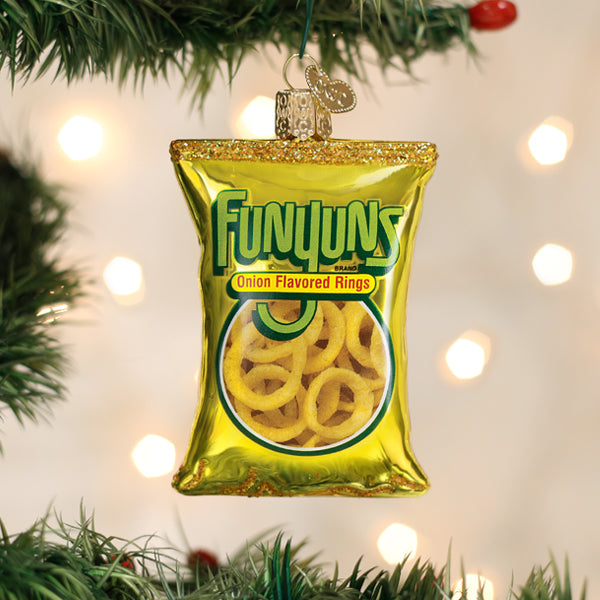 A Christmas tree ornament shaped like a shiny yellow bag of Funyuns onion flavored rings hangs among green pine branches with blurred lights in the background.