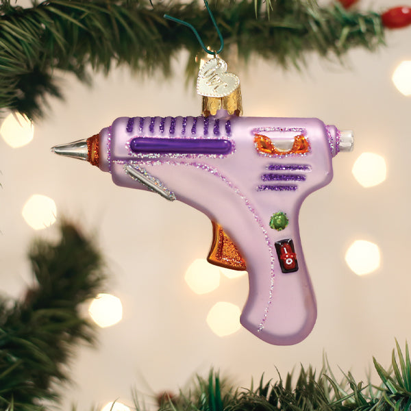 A glittery, purple hot glue gun ornament hangs on a Christmas tree branch, with blurred warm lights in the background.