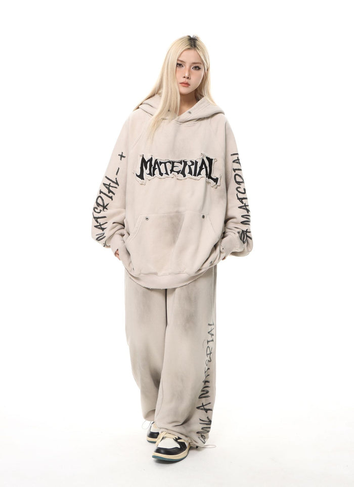 Front model view of a washed beige oversized hoodie with chest lettering, printed sleeves, and matching wide-leg pants
