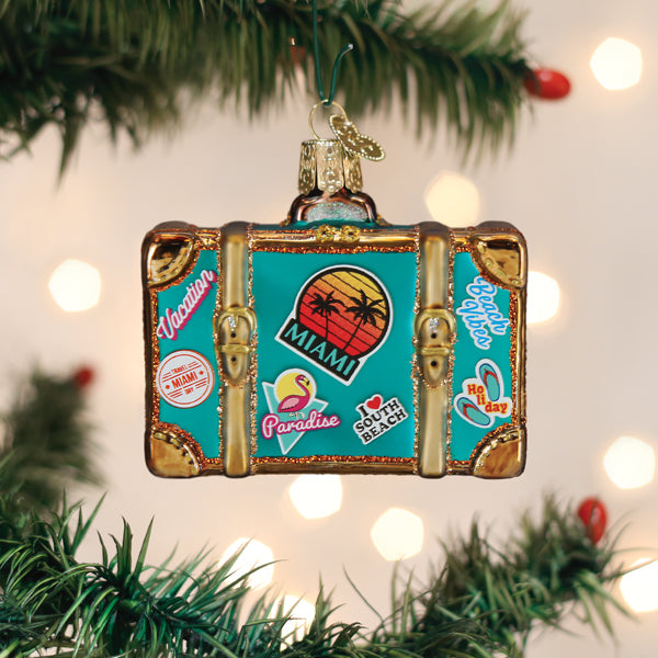 A suitcase-shaped ornament decorated with colorful Miami-themed stickers hangs on a Christmas tree, with out-of-focus lights twinkling softly in the background.
