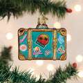 A suitcase-shaped ornament decorated with colorful Miami-themed stickers hangs on a Christmas tree, with out-of-focus lights twinkling softly in the background.