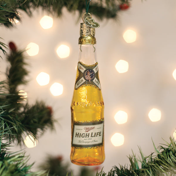 A glass ornament shaped like a Miller High Life beer bottle hangs on a Christmas tree, surrounded by green branches and blurred white holiday lights in the background.