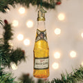A glass ornament shaped like a Miller High Life beer bottle hangs on a Christmas tree, surrounded by green branches and blurred white holiday lights in the background.