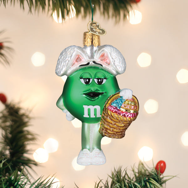 A green M&M ornament dressed with white bunny ears and shoes, holding a basket of colorful eggs, hangs on a Christmas tree with blurred lights in the background.