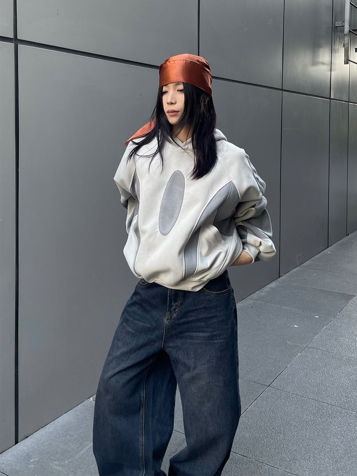 Model wearing an oversized light gray hoodie with darker gray patchwork panels and oval front detail