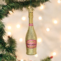 A glittery gold and pink prosecco bottle ornament hangs on a Christmas tree, with blurred white holiday lights in the background.