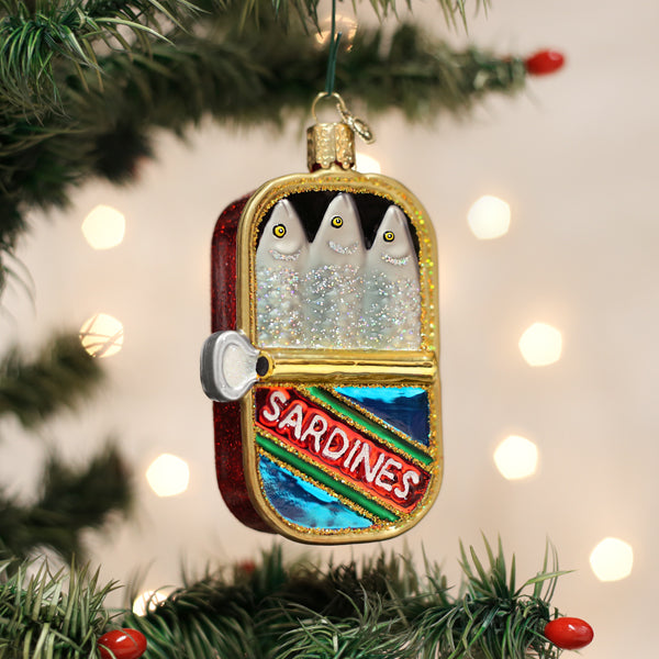 A glittery Christmas tree ornament shaped like an open tin of sardines hangs on a tree branch. The tin has three cartoon-like fish inside and a label that reads SARDINES. Blurred lights are in the background.