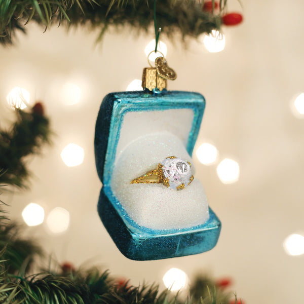 A gold ring with a large clear gemstone sits in a blue velvet ring box ornament, hanging on a Christmas tree with blurred white lights in the background.