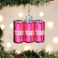 A Christmas ornament shaped like three pink Tab soda cans hangs on a pine tree branch, with soft white holiday lights blurred in the background.